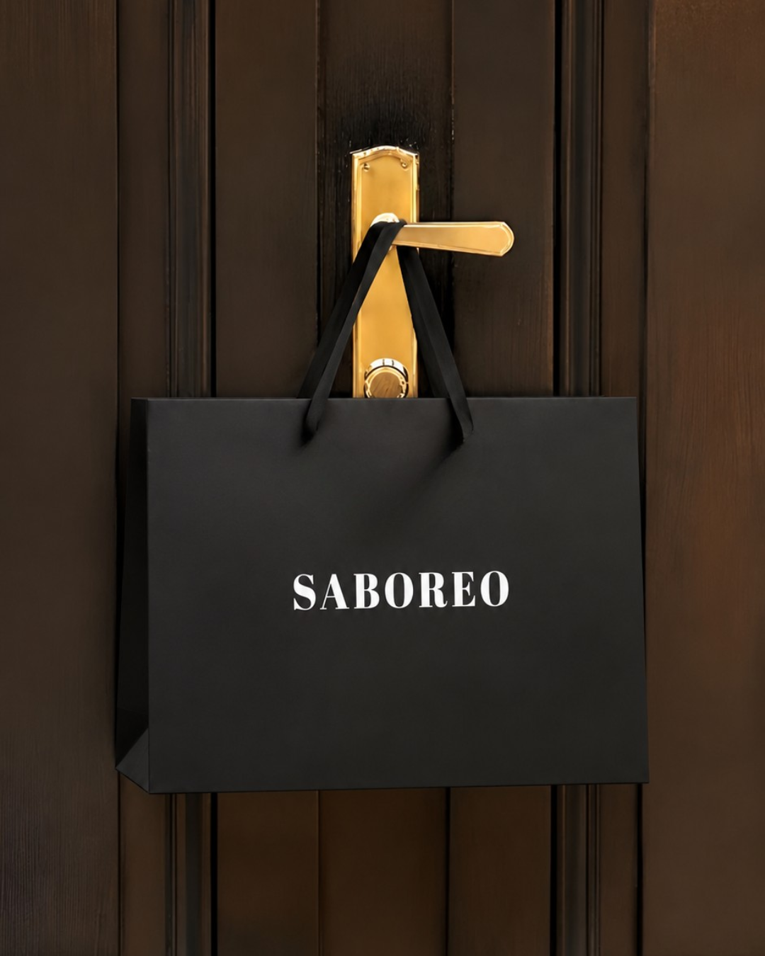 A black Saboreo gift bag hanging from a brass door handle on a dark wooden door
